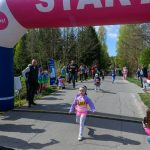 Dziewczynka w fioletowym stroju biegnie pod różowym łukiem startowym. W tle las i ludzie wiwatujący. Dzień jest słoneczny.