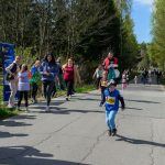 Dziecko biegnie w wyścigu, otoczone osobami kibicującymi. W tle widać drzewa i niebo. Na przodzie numer startowy 286.