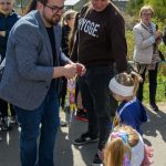 Na zdjęciu grupa osób na zewnątrz, mężczyzna w okularach wręcza medale dzieciom. Tło to zielone drzewa i jasne niebo. Dzieci mają kolorowe ubrania i wstążki.