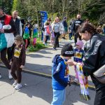 Na zdjęciu dzieci biorą udział w biegu na zewnątrz. Osoba wręcza medale, jest słoneczny dzień, wokół są inni uczestnicy i drzewa.