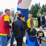 Grupa ludzi w kolorowych ubraniach stoi obok dużego dmuchanego łuku. W tle widać drzewa. Jasny dzień na zewnątrz.