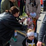 Dziecko w numerze startowym 20 uśmiecha się, trzymając za rękę dorosłego. W tle ludzie na zewnątrz, drzewa i niebie. Dzieci noszą kolorowe czapki.