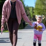 Na zdjęciu kobieta i dziecko idą za rękę w słoneczny dzień. Kobieta nosi różową kurtkę, dziecko ma numerek startowy. Tło to zielone drzewa i niebo.