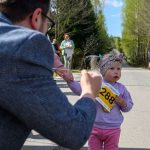 Małe dziecko z numerem startowym 288 na piersi jest trzymane za rękę przez dorosłego. Ubrana jest w różowo-fioletowe ubranko. W tle widać las i kilka osób na drodze.