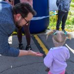 Mężczyzna w okularach pochyla się ku małej dziewczynce z kucykami. Ona ubrana jest w różowy sweterek. W tle widać asfalt i trawę.