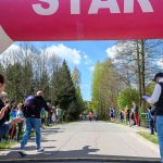 Na zdjęciu widać start biegowy z wielkim, różowym balonem z napisem START. Zawodnicy biegną po asfaltowej drodze, a widzowie stoją przy trasie wśród drzew. Tło stanowi niebo z białymi chmurami.