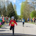 Dwóch dzieci w kolorowych strojach biegnie wyścig. W tle las, a po bokach kibicujący ludzie. Jasny, słoneczny dzień.