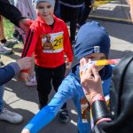 Dzieci w kolorowych ubraniach uczestniczą w biegu na zewnątrz. Jedno z nich ma numer startowy, a kobieta wręcza mu pamiątkowy medal. Tło stanowią inni uczestnicy i fragment zieleni.