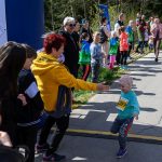 Małe dziecko z numerem startowym biegnie do mety na asfaltowej drodze. W tle grupa kibicujących osób, drzewa i niebieski banner. Dzień jest słoneczny, a atmosfera radosna.