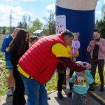 Grupa osób na świeżym powietrzu, mężczyzna w czerwonej kamizelce wręcza medale dzieciom. Tło to zielony krajobraz z niebieskim niebem. Uczestnicy maja na sobie kolorowe ubrania, a atmosfera jest radosna.