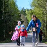 Na zdjęciu widać trzy osoby spacerujące po ścieżce otoczonej drzewami. Osoba dorosła ubrana w niebieską kurtkę trzyma smartfon. Dwoje dzieci ubranych w kolorowe kurtki idzie obok.