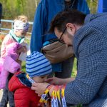 Na zdjęciu osoba zakłada medal dziecku w czerwonej kurtce. W tle dziecko w różowej kurtce i dorosły w dżinsowej koszuli. Otoczenie jest zielone, sugerujące wiosenny plener.