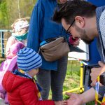 Na zdjęciu jest małe dziecko w czerwonej kurtce, które ściska dłoń dorosłego. W tle są uśmiechnięta kobieta i inne dziecko. Otoczenie to zielone drzewa i niebieskie niebo.