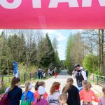 Na zdjęciu grupa dzieci stoi pod różowym banerem START. W tle widoczna jest droga w otoczeniu drzew. Pogoda jest słoneczna, a przy drodze stoi kilka osób.