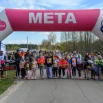 Dzieci stoją na linii startu pod różowo-niebieskim łukiem z napisem META. Tło stanowi zieleń i zebrani ludzie. Słoneczny dzień, jasne kolory.