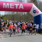 Dzieci w kolorowych strojach startują w biegu pod różowym łukiem z napisem META. W tle las i zgromadzeni kibice.