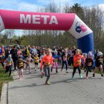 Grupa dzieci biegnie pod różowo-białym łukiem z napisem META. W tle liczny tłum na tle drzew. Dzień jest słoneczny.