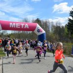 Dzieci biegną przez różowo-białą bramkę z napisem META. Tło stanowi las i niebieskie niebo z chmurami. Uczestnicy mają na sobie kolorowe stroje i numery startowe.