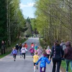 Grupa dzieci biegnie drogą w lesie, otoczona jest przez dorosłych. Drzewa są zielone, a niebo jest błękitne. Kilku dorosłych robi zdjęcia.