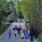 Dzieci biegają po leśnej drodze, otoczone zielonymi drzewami. Tło stanowi wiosenny las. Ubrania dzieci są kolorowe.