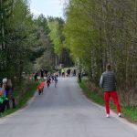 Bieg dzieci na wiejskiej drodze między drzewami wiosną. W tle kibicujący dorośli, dzieci w kolorowych ubraniach. Pogoda jasna, pochmurna.