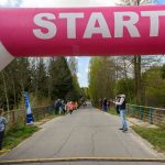 Na zdjęciu widać różowy balon startowy na biegu, pod nim biegną dzieci. W tle zieleń drzew i niebo z chmurami. Ludzie stoją wzdłuż trasy.