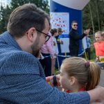 Mężczyzna w marynarce rozmawia z dziewczynką w stroju sportowym na zawodach. W tle ludzie oraz balon z napisem Ale czas!. Scena odbywa się na zewnątrz, w pobliżu drzew.