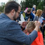 Mężczyzna w szarej marynarce zakłada medal dziecku w pomarańczowej bluzie. W tle widać drzewa i grupę ludzi. Dziecko ma numer startowy 204.