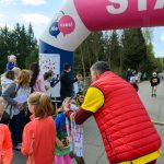 Na zdjęciu widoczna jest ceremonia wręczenia medali ukończenia biegu dzieciom przy różowo-białym łuku startowym na tle lasu. Tłum ludzi w kolorowych ubraniach obserwuje wydarzenie. Atmosfera jest radosna, a otoczenie zielone.