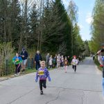 Dzieci biegają w kolorowych strojach na wiejskiej drodze, otoczonej drzewami. Stojący mężczyzna z mikrofonem prowadzi wydarzenie. Baner reklamowy widoczny z lewej strony drogi.