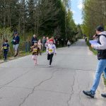 Na zdjęciu widać bieg dzieci na wiejskiej drodze, w tle las. Baner i kibice po lewej, komentator z mikrofonem po prawej. Dzieci mają numery startowe.