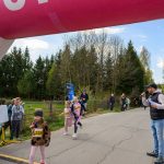 Dzieci biegną pod różowym łukiem z numerami startowymi na jasnej drodze. W tle drzewa i widzowie. Mężczyzna z mikrofonem kibicuje.