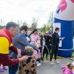 Na zdjęciu grupa osób przy starcie biegu, ubrani kolorowo. Tło to zielone drzewa, niebo zachmurzone. Widać dużą, niebiesko-różową bramę z napisem.