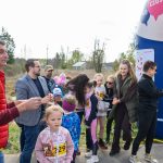 Grupa ludzi, w tym dzieci i dorośli, stoi na zewnątrz obok niebieskiego dmuchanego łuku. Tło stanowi zalesiony teren, wszyscy wyglądają na radosnych. Uczestnicy noszą numery startowe, co sugeruje udział w wydarzeniu sportowym