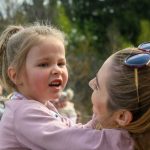 Dziewczynka w różowej kurtce trzymana przez dorosłą osobę na tle rozmytego zielonego krajobrazu. Z przodu widać także kobietę w okularach przeciwsłonecznych. Otoczenie jest zewnętrzne, naturalne.