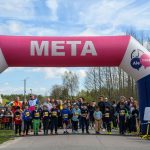 Ludzie gromadzą się na starcie biegu pod różowo-białym łukiem META. Dzieci w sportowych strojach z numerami na piersi. W tle zielone drzewa i błękitne niebo.