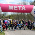 Rozpoczęcie biegu dzieci na asfaltowej drodze pod różowym balonem z napisem META. Tło to zieleń i drzewa wiosną. Wokół kibice i rodzice.
