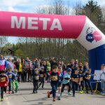 Dzieci biegają pod różowo-niebieskim łukiem z napisem META. Na tle widać liczną publiczność oraz drzewa. Pogoda jest słoneczna.