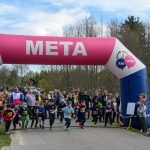 Bieg dzieci zaczyna się pod różowo-niebieskim łukiem z napisem META. W tle las i niebo z chmurami. Po bokach kibice.