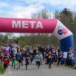 Dzieci biegną pod różowo-niebieskim łukiem z napisem META. W tle drzewa i niebo. Obok zgromadzeni kibice.