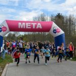 Bieg dzieci pod różowo-niebieską bramą z napisem META. Tło stanowi widownia na tle drzew i błękitnego nieba. Dzieci w kolorowych strojach biegną po kostce brukowej.