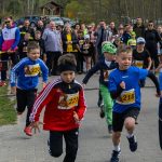 Młodzi biegacze startują w wyścigu na tle licznej publiczności. Kolorowe stroje dzieci kontrastują z zielenią w tle. Atmosfera sportowego wydarzenia z licznikiem czasowym.