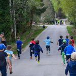 Dzieci biegną wyścigiem po asfaltowej drodze otoczonej zielonym lasem. Po lewej stronie kilku dorosłych obserwuje wydarzenie. Na końcu drogi widoczni są organizatorzy w kamizelkach.