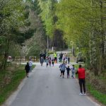 Droga w lesie z grupą biegnących dzieci i dorosłych. Zielone drzewa i niebo w tle. Ludzie ubrani w kolorowe stroje.