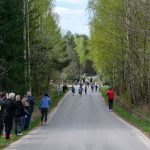 Na zdjęciu bieg uliczny dzieci na wiejskiej drodze. Wokół zielone drzewa i kibice. Pogoda jest pochmurna.