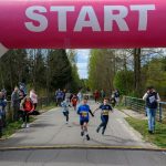 Dzieci biegną pod różowym łukiem START na leśnej drodze. W tle kibice i drzewa. Na ziemi widoczny jest zegar czasu 0:00:52.