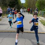 Dzieci w kolorowych koszulkach biegną ulicą podczas wyścigu. W tle zieleń i kibice przy barierkach dopingują. Na skraju mężczyzna z mikrofonem.