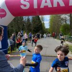 Dzieci przekraczają linię startową na zawodach biegowych. Tło stanowi zielony las. Nad nimi znajduje się różowy baner „START”.