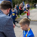 Na zdjęciu widać grupkę dzieci biegnących alejką. Chłopiec w niebieskiej koszulce z medalem stoi z przodu. W tle ludzie obserwują bieg pośród drzew.
