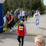 Uczestnicy biegu na szosie, głównie dzieci, otoczeni drzewami. Żółte numery startowe, ubrania w odcieniach czerwieni i bieli. W tle kibicujący ludzie i niebieski balon startowy.